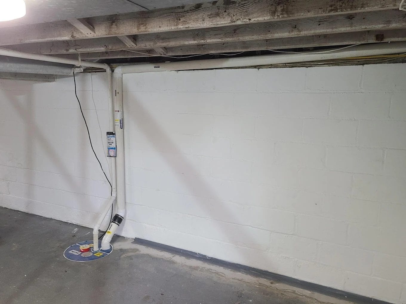 White piping for radon mitigation that's moving down a wall and into the flooring of a basement.